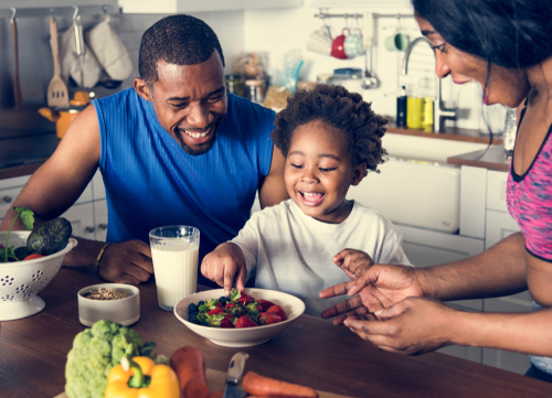 Two parents smiling as their young daughter eats strawberries from a bowl