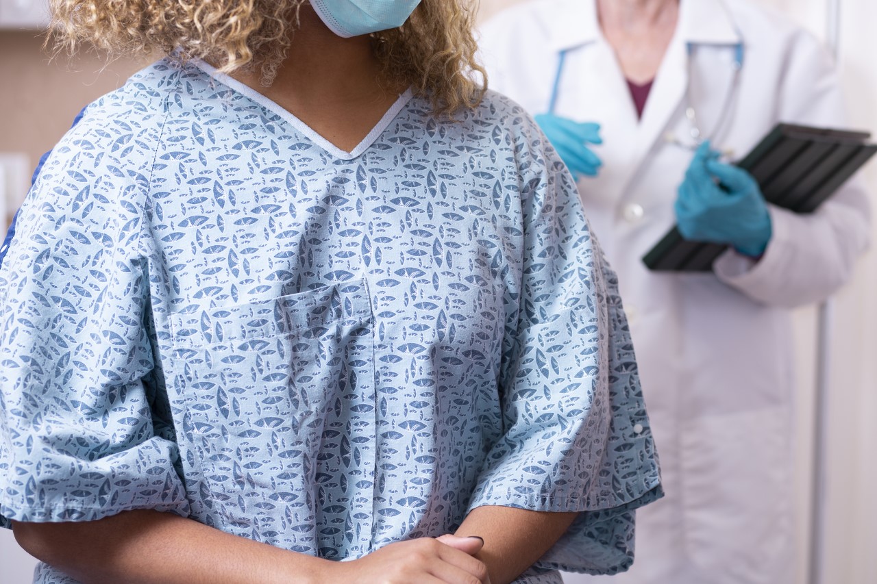Closeup of a woman in a blue hospital gown, preparing for a mammogram while an oncologist stands behind her