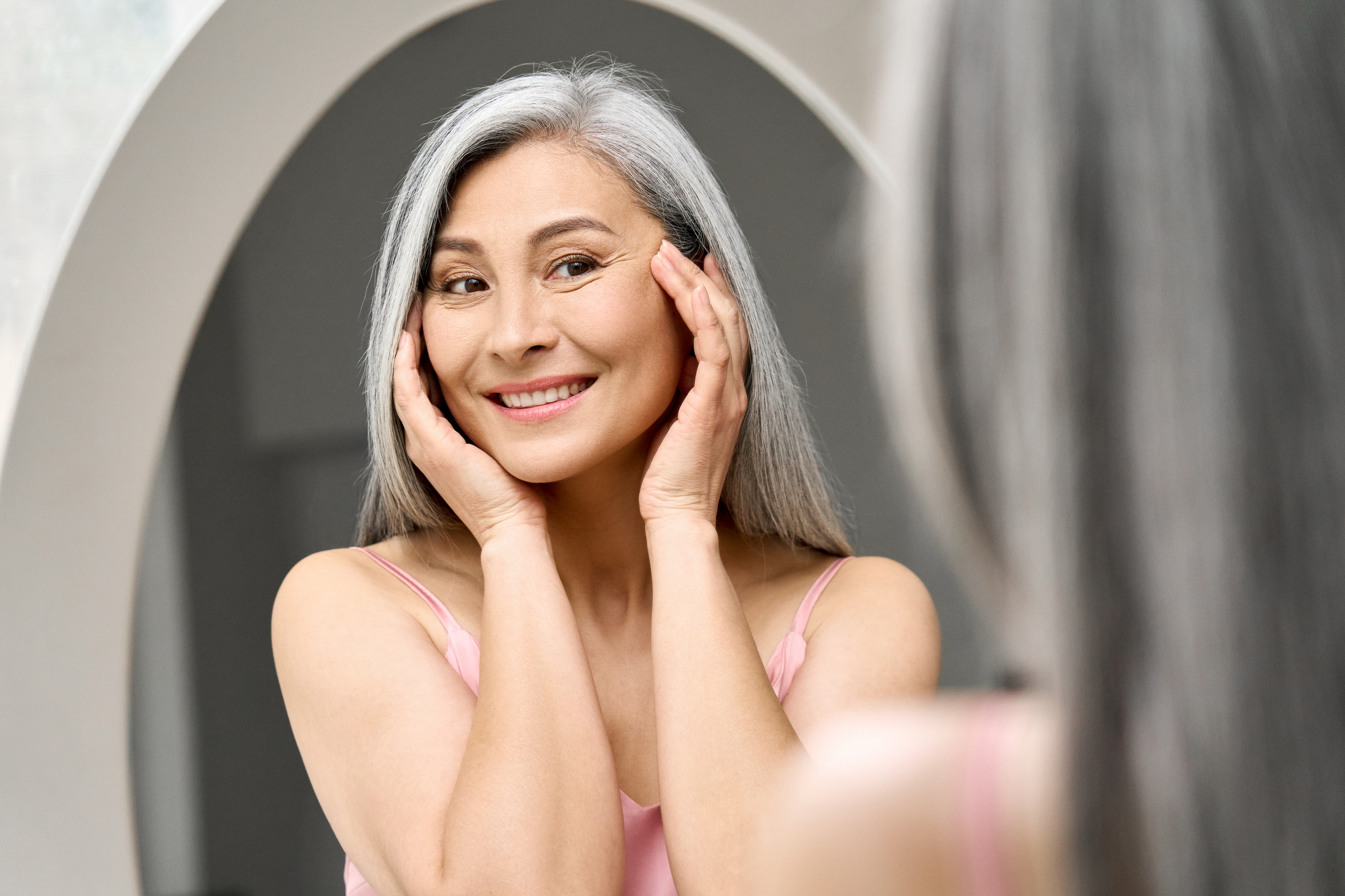 Grey-haired woman smiling and examining her face in a mirror after a successful cosmetic neurotoxin treatment