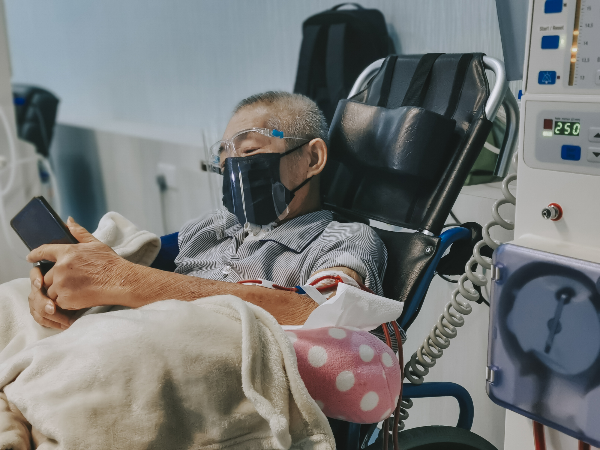 Masked patient looking at his phone while undergoing the kidney dialysis process