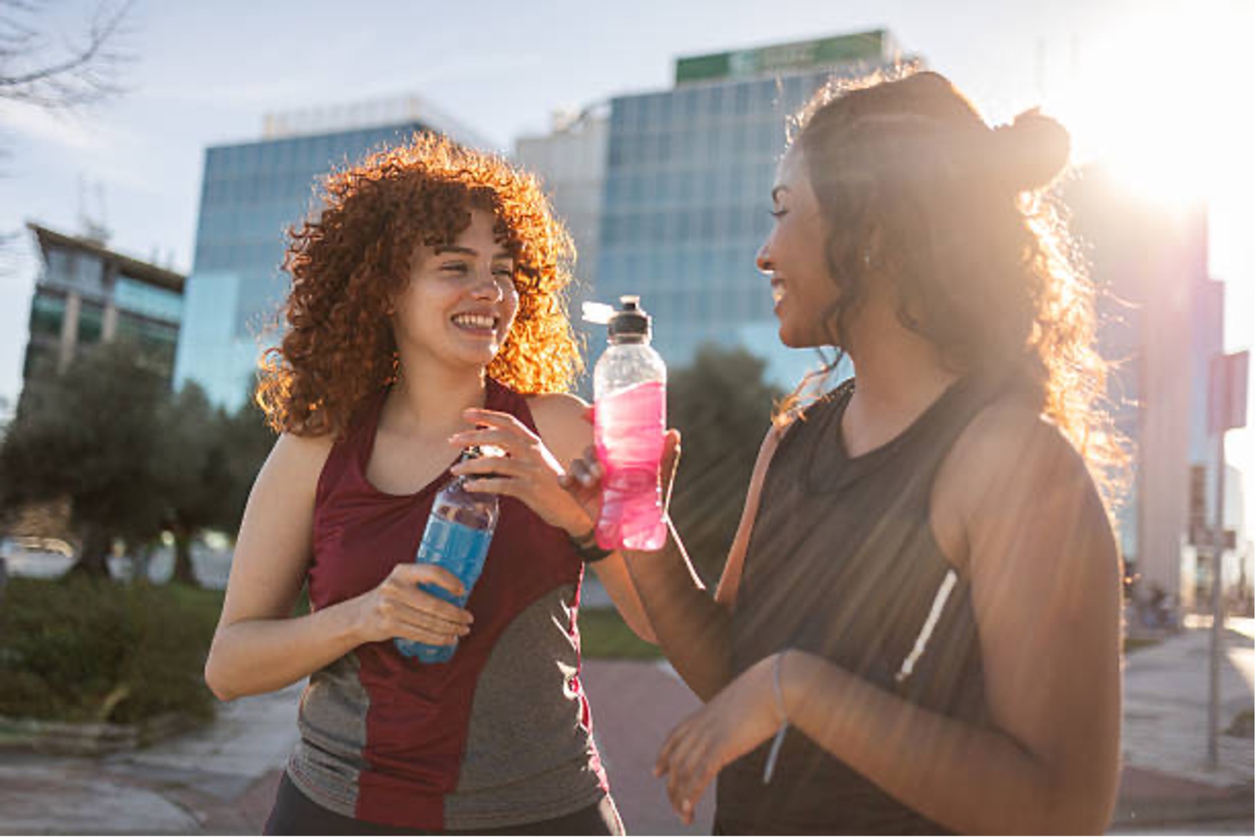 Two multi-ethnic female athletes taking a break from their workout, enjoying refreshing water, and chatting happily on a sunny day in an urban environment filled with modern buildings