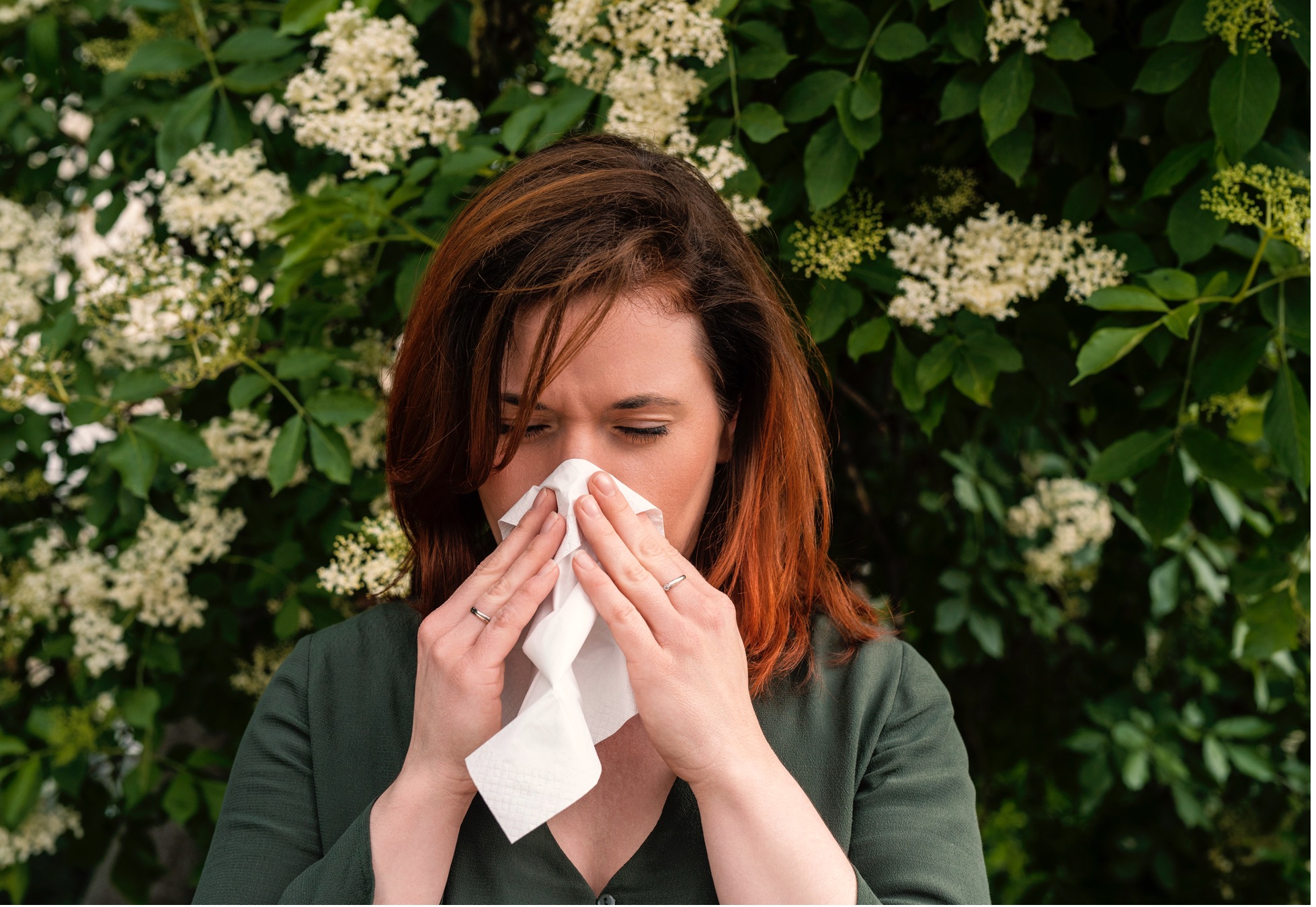 A woman sneezing outdoors surrounded by blooming flowers, representing pollen allergies in the spring.