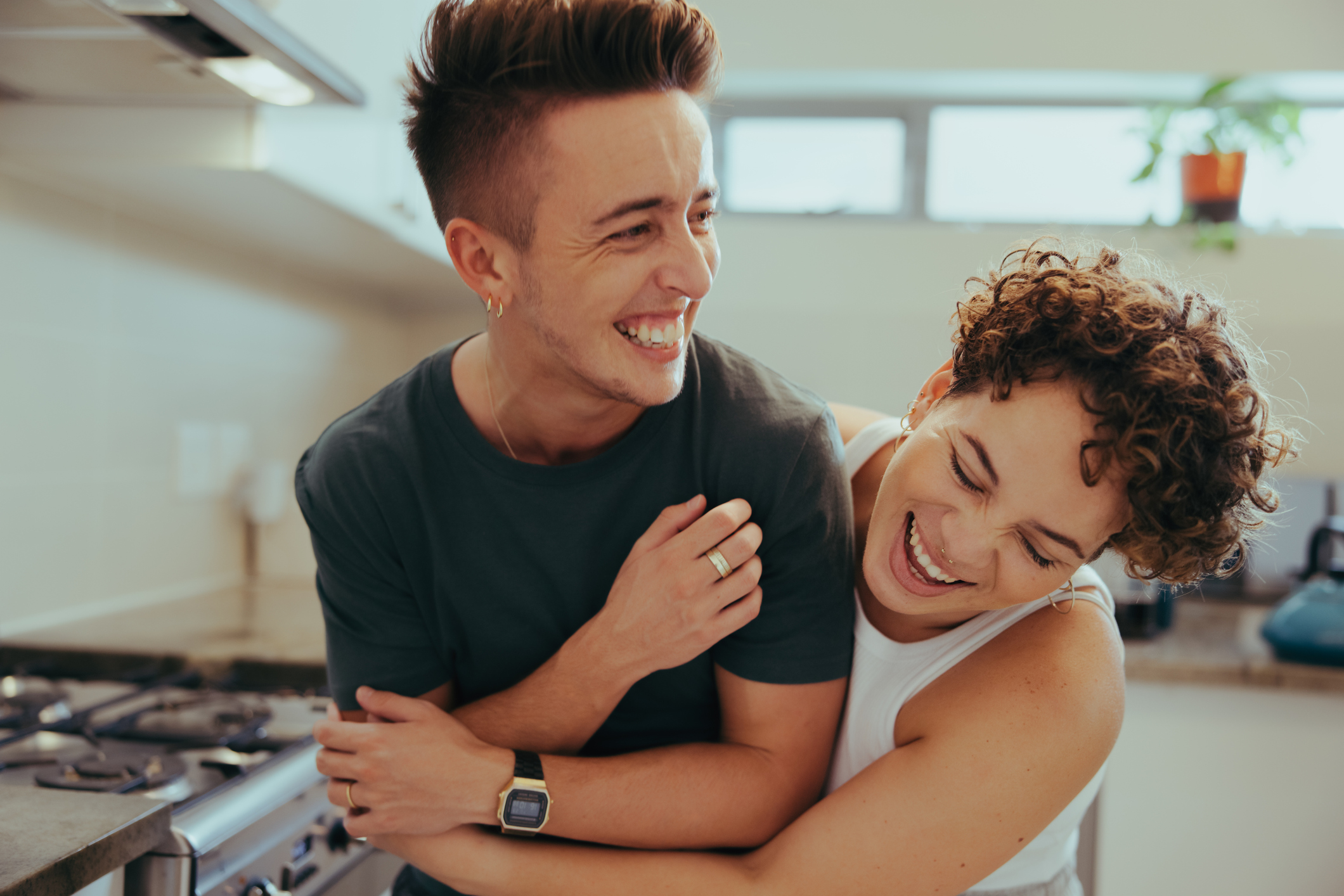 Young queer couple laughing together indoors. Happy young queer couple having fun together while standing in their kitchen. Romantic young LGBTQ+ couple bonding fondly at home.
