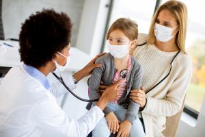 Masked doctor using a stethoscope on a young girl during an annual physical