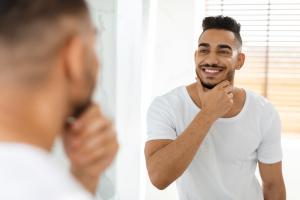 Male Facial Care. Handsome Arab Man Looking In Mirror In Bathroom And Touching Beard, Attractive Masculine Middle Eastern Guy Smiling To Reflection, Enjoying His Appearance, Selective Focus