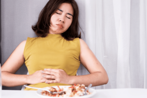 Woman in yellow top holding stomach showing discomfort from bloating after eating a meal