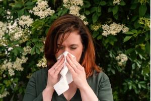 A woman sneezing outdoors surrounded by blooming flowers, representing pollen allergies in the spring.