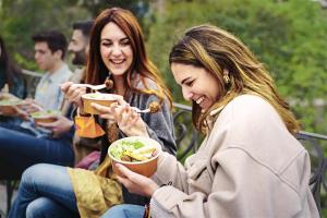 Young people gather and eat salads while enjoying the outdoors together in the park.