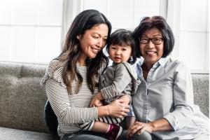 Mother and grandmother smiling while holding a young child on a couch, representing multigenerational women’s health and menopause care and support